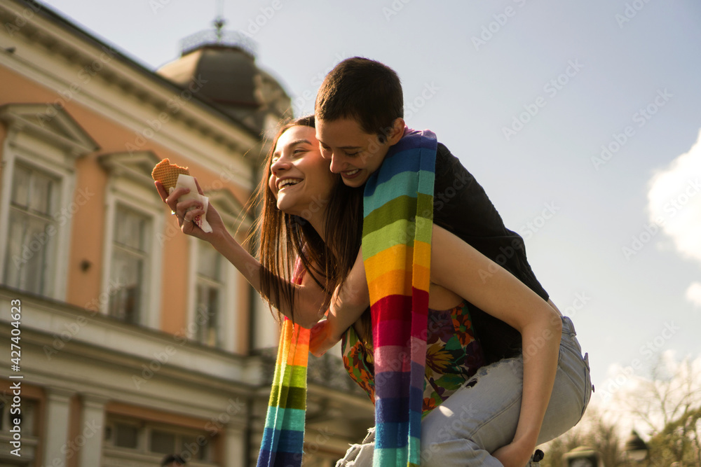 A young lesbian couple embraces their love. They are walking the streets and eating ice cream covered together with a rainbow colors scarf.