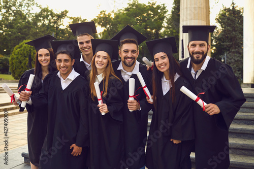 Group of happy young people university graduates in traditional bonets and masters mantles walking with diplomas in raised hands and looking at camera over university building background, side view
