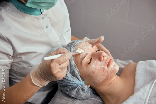 A female cosmetologist in a medical mask applies cream to the client's face with a brush. Hydration and rejuvenation of the skin. Professional skin care.