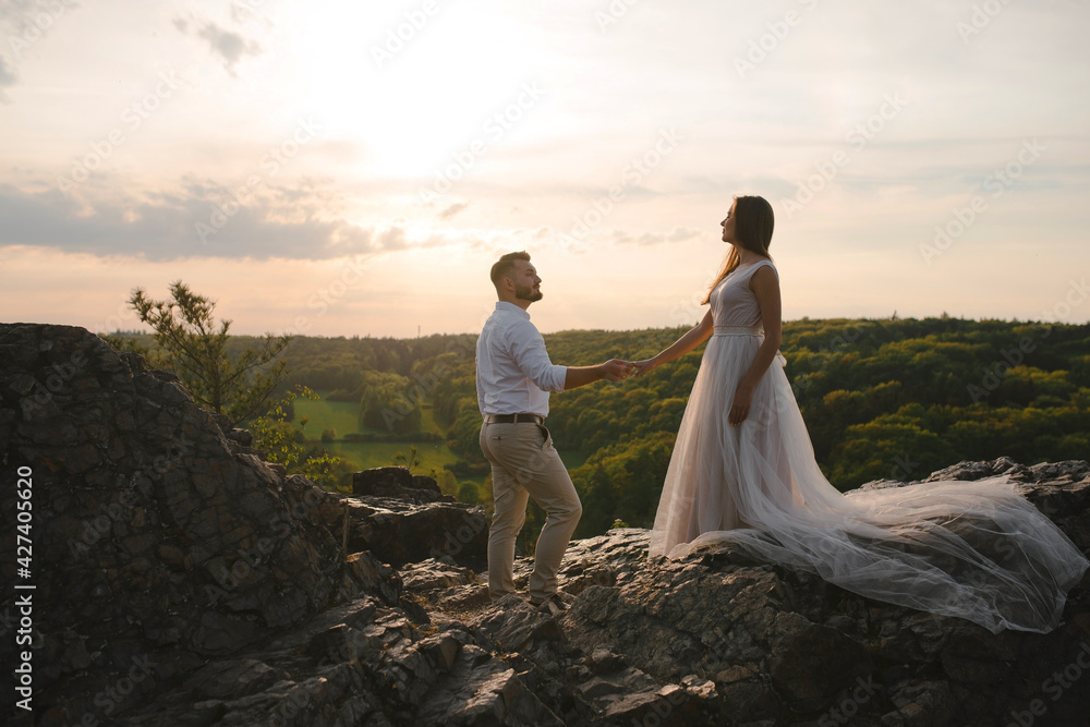Romantic wedding couple standing on the rocks on sunset. Bride woman in beautiful dress with long loop and stylish groom holding hands. Sunset in green mountains Romantic wedding couple standing on the rocks on sunset. Bride woman in beautiful dress with long loop and stylish groom holding hands. Sunset in green mountains