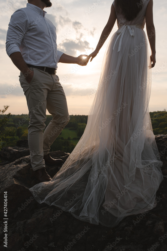 Rear view shot of romantic wedding couple standing on the rocks on sunset. Bride woman in beautiful dress with long loop and stylish groom holding hands. Sunset in mountains Rear view shot of romantic wedding couple standing on the rocks on sunset. Bride woman in beautiful dress with long loop and stylish groom holding hands. Sunset in mountains
