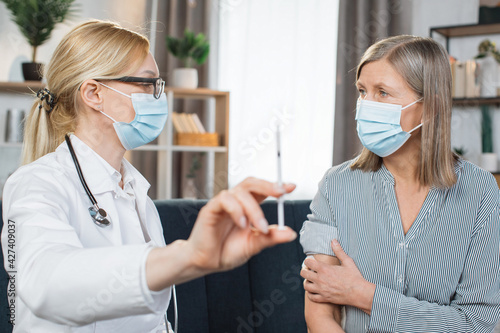 Close up of likable experienced mature woman doctor in protective mask  showing syringe at camera  talking with her senior female patient before injection of vaccine or insuline during home visit.