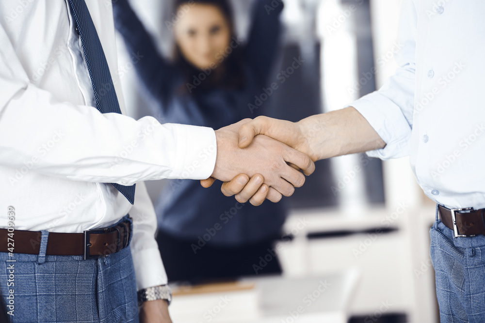 Fototapeta premium Two businessmen are shaking hands in office, close-up. Happy and excited business woman stands with raising hands at the background. Business people concept