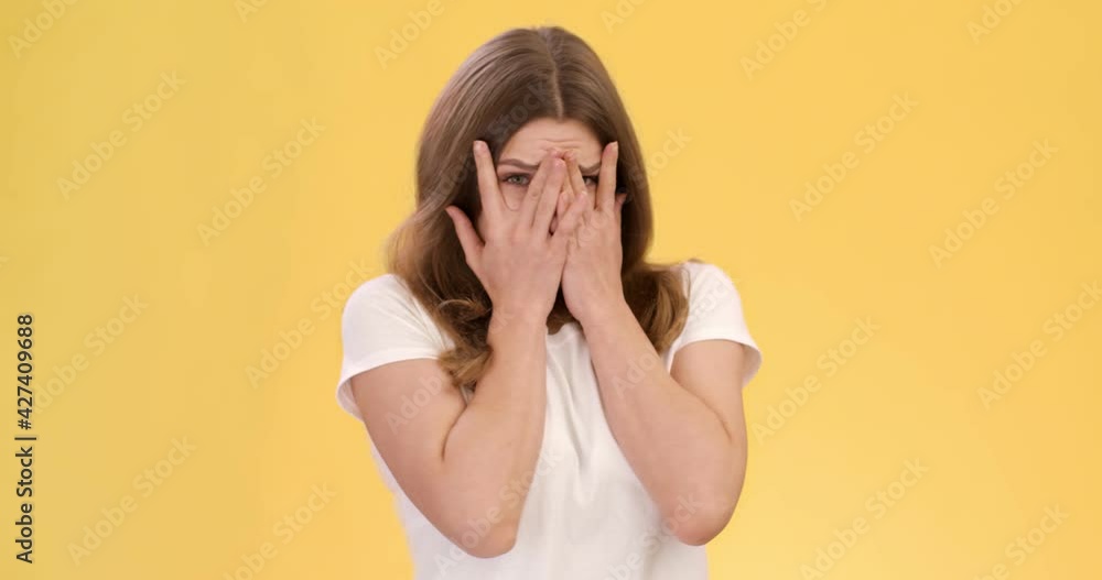 Young afraid woman feeling scared, covering her face with hands and peeking through fingers, orange studio background Young afraid woman feeling scared, covering her face with hands and peeking through fingers, orange studio background