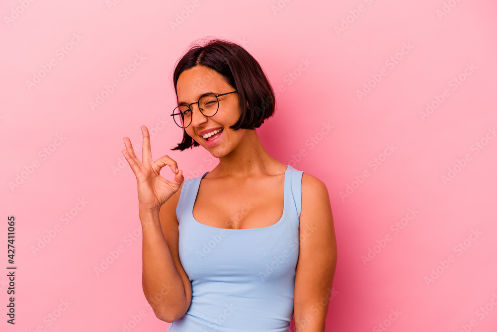 Young mixed race woman isolated on pink background winks an eye and holds an okay gesture with hand. Young mixed race woman isolated on pink background winks an eye and holds an okay gesture with hand.