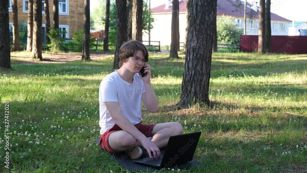 Young man is sitting on the grass in park with his laptop and talking on the phone. Distance learning concept. 4k footage Young man is sitting on the grass in park with his laptop and talking on the phone. Distance learning concept. 4k footage