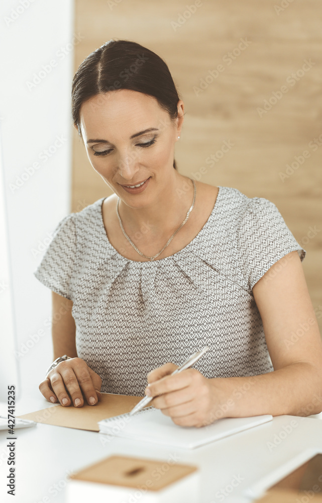 Cheerful smiling businesswoman working with computer while sitting at the desk in modern office. Middle aged female lawyer or auditor at work. Business people concept Cheerful smiling businesswoman working with computer while sitting at the desk in modern office. Middle aged female lawyer or auditor at work. Business people concept
