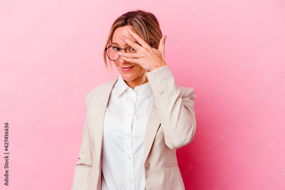Young caucasian mixed race business woman isolated on pink background blink at the camera through fingers, embarrassed covering face.