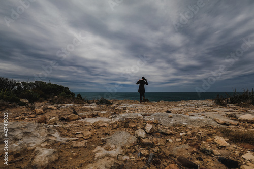 Landscape image featuring woman standing on rocky cliff looking out at sea