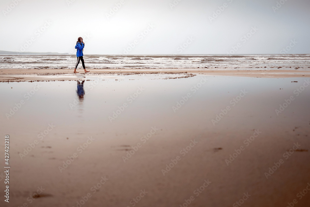 Teenager girl walking on a sandy beach. The model dressed in blue jacket and black leggins. Outdoor activity concept. Selective focus. Warm tone