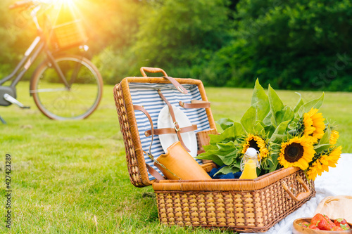 Summer picnic concept on sunny day with watermelon, fruit, bouquet hydrangea and sunflowers flowers. Picnic basket on grass with food and refreshing summer drink on white knit blanket. Selective focus
