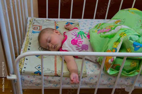 A small child sleeps, covered with a blanket, in a white metal bed in a hospital or orphanage photo