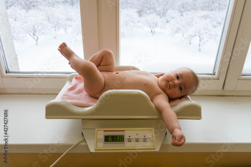Weighing a baby: a small naked baby lies on a pink diaper-covered newborn electronic scale in a hospital on a white windowsill