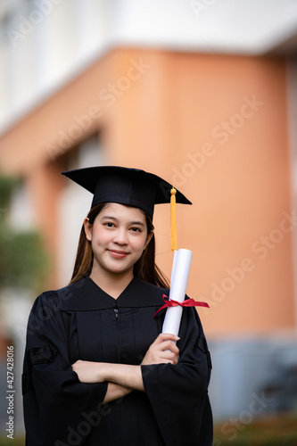 A young happy Asian woman university graduate in graduation gown and mortarboard holds a degree certificate celebrates education achievement in the university campus. Education stock photo