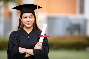 A young happy Asian woman university graduate in graduation gown and mortarboard holds a degree certificate celebrates education achievement in the university campus.  Education stock photo