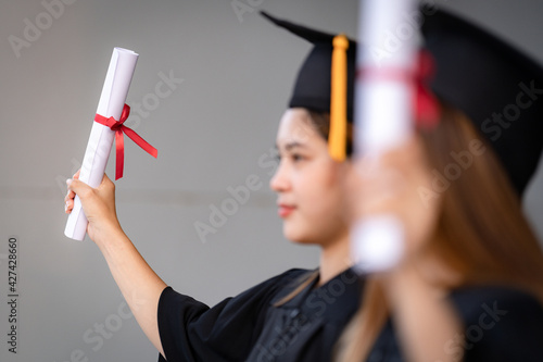 A young happy Asian woman university graduate in graduation gown and mortarboard holds a degree certificate celebrates education achievement in the university campus. Education stock photo