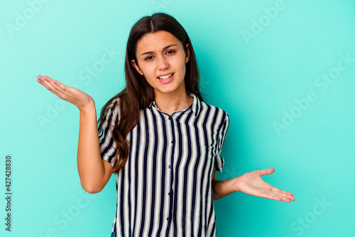 Young Indian woman isolated on blue background confused and doubtful shrugging shoulders to hold a copy space.
