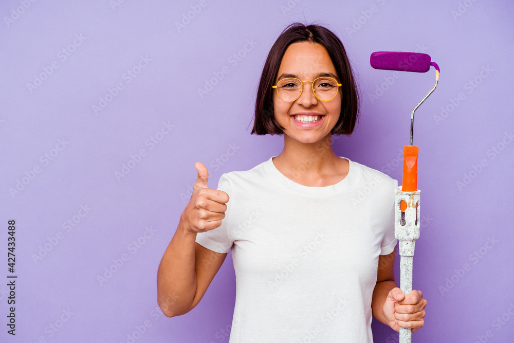 Young painter mixed race woman holding a paint stick isolated on purple background smiling and raising thumb up Young painter mixed race woman holding a paint stick isolated on purple background smiling and raising thumb up