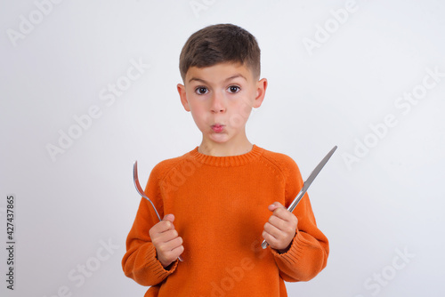 Model hungry Caucasian kid boy wearing knitted sweater against white wall holding in hand fork knife want tasty yummy pizza pie