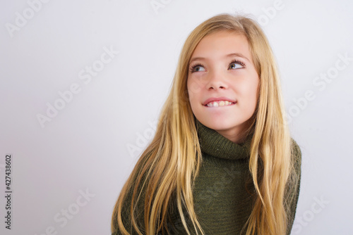 Amazed Caucasian kid girl wearing green knitted sweater against white wall bitting lip and looking tricky to empty space.