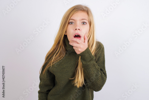 Nervous puzzled Caucasian kid girl wearing green knitted sweater against white wall opens mouth from surprise, reacts on sudden news.