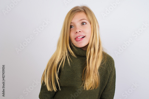 Caucasian kid girl wearing green knitted sweater against white wall showing grimace face crossing eyes and showing tongue. Being funny and crazy