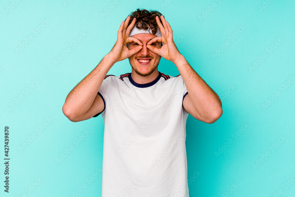 Young sport caucasian man isolated on blue background showing okay sign over eyes Young sport caucasian man isolated on blue background showing okay sign over eyes