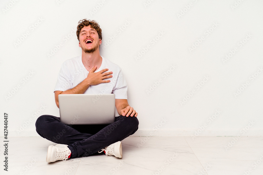 Young caucasian man sitting on the floor holding on laptop isolated on white background laughs out loudly keeping hand on chest. Young caucasian man sitting on the floor holding on laptop isolated on white background laughs out loudly keeping hand on chest.