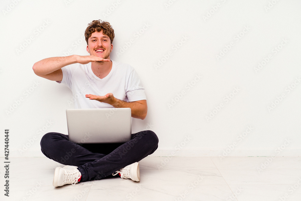 Young caucasian man sitting on the floor holding on laptop isolated on white background holding something with both hands, product presentation. Young caucasian man sitting on the floor holding on laptop isolated on white background holding something with both hands, product presentation.