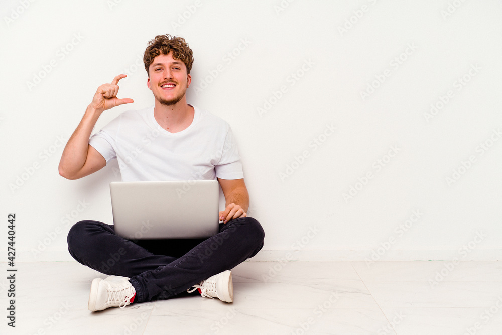 Young caucasian man sitting on the floor holding on laptop isolated on white background holding something little with forefingers, smiling and confident. Young caucasian man sitting on the floor holding on laptop isolated on white background holding something little with forefingers, smiling and confident.