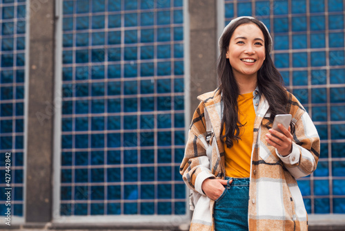 A beautiful young Asian woman is holding her phone and smiles in front of a blue tiled wall, wearing yellow t-shirt, jeans and beret. 