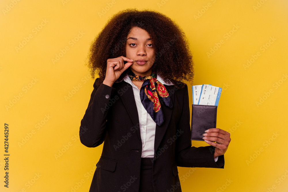 Young african american air hostess holding a plane tickets isolated on yellow background with fingers on lips keeping a secret. Young african american air hostess holding a plane tickets isolated on yellow background with fingers on lips keeping a secret.