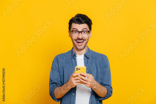 Happy handsome amazed stylish guy with eyeglasses, using smartphone, chatting online, writing message, browsing internet, social media, looks surprised at camera, standing on isolated orange photo