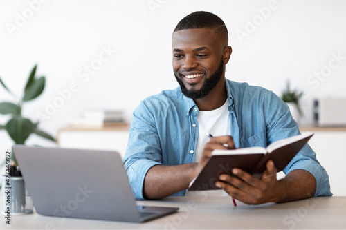 Black Millennial Freelancer Guy Taking Notes While Working On Laptop At Home