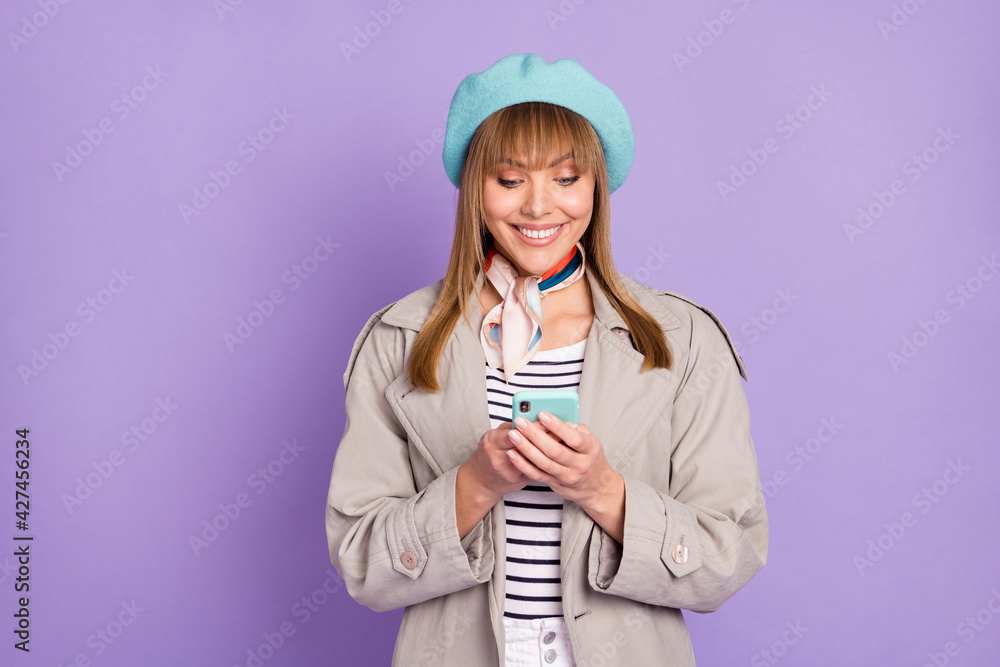 Photo of cute girl hold telephone look screen white smile wear blue beret scarf coat isolated purple color background Photo of cute girl hold telephone look screen white smile wear blue beret scarf coat isolated purple color background
