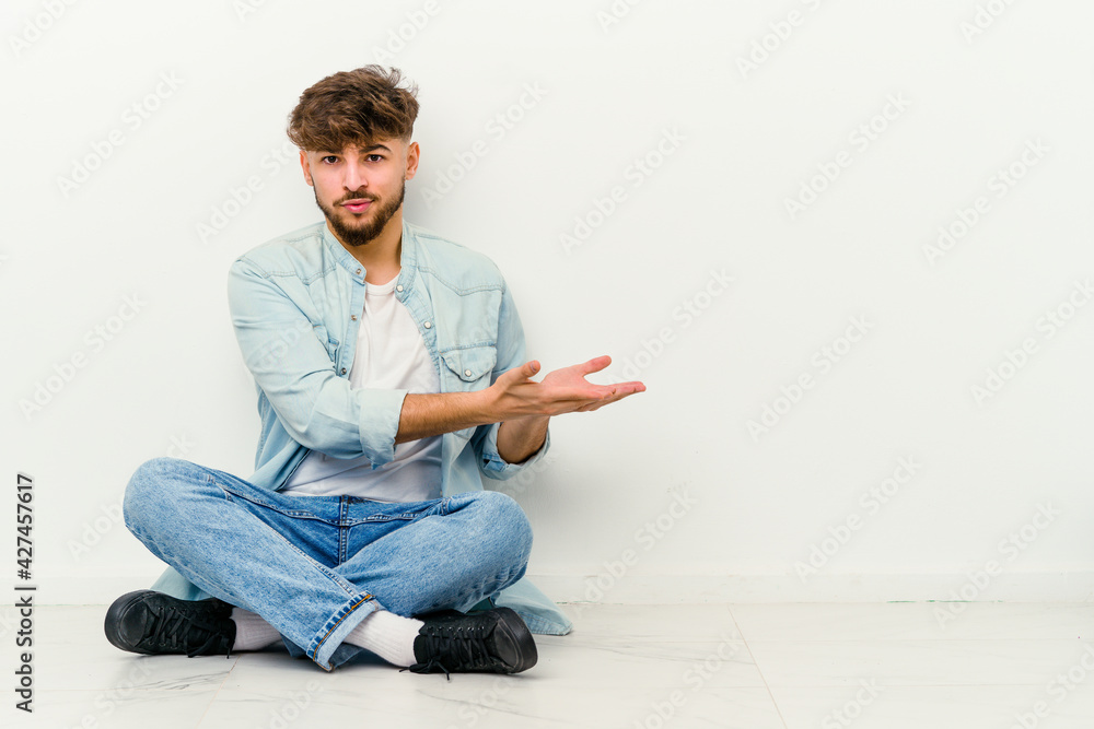 Young Moroccan man sitting on the floor isolated on white background holding a copy space on a palm. Young Moroccan man sitting on the floor isolated on white background holding a copy space on a palm.