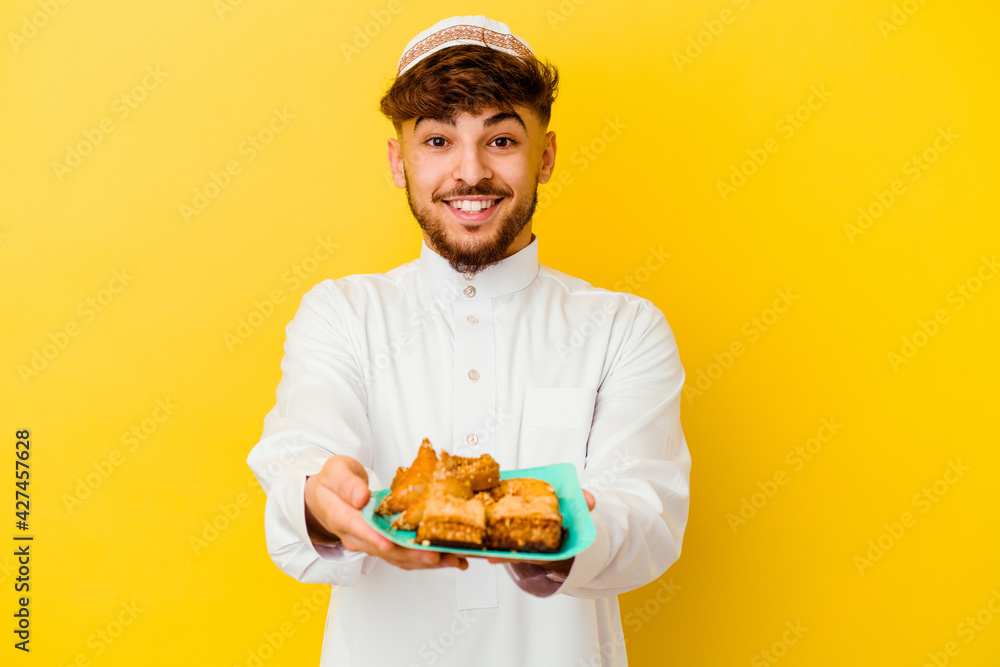 Young Moroccan man wearing the typical arabic costume eating Arabian sweets isolated on yellow background Young Moroccan man wearing the typical arabic costume eating Arabian sweets isolated on yellow background