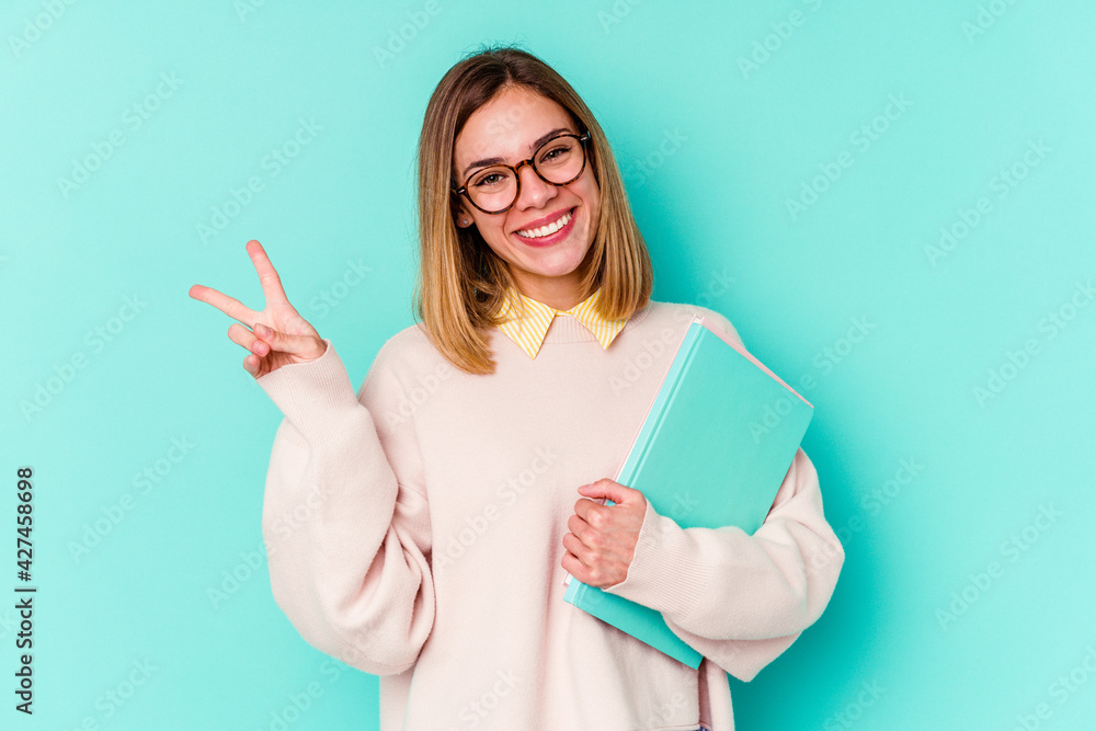 Young student woman holding books isolated on blue background joyful and carefree showing a peace symbol with fingers. Young student woman holding books isolated on blue background joyful and carefree showing a peace symbol with fingers.