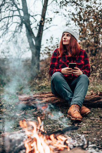 Traveler girl in hipster clothes sits on a log enjoying, relaxing in the forest near the fire by camping. Woman using smartphone in nature photo