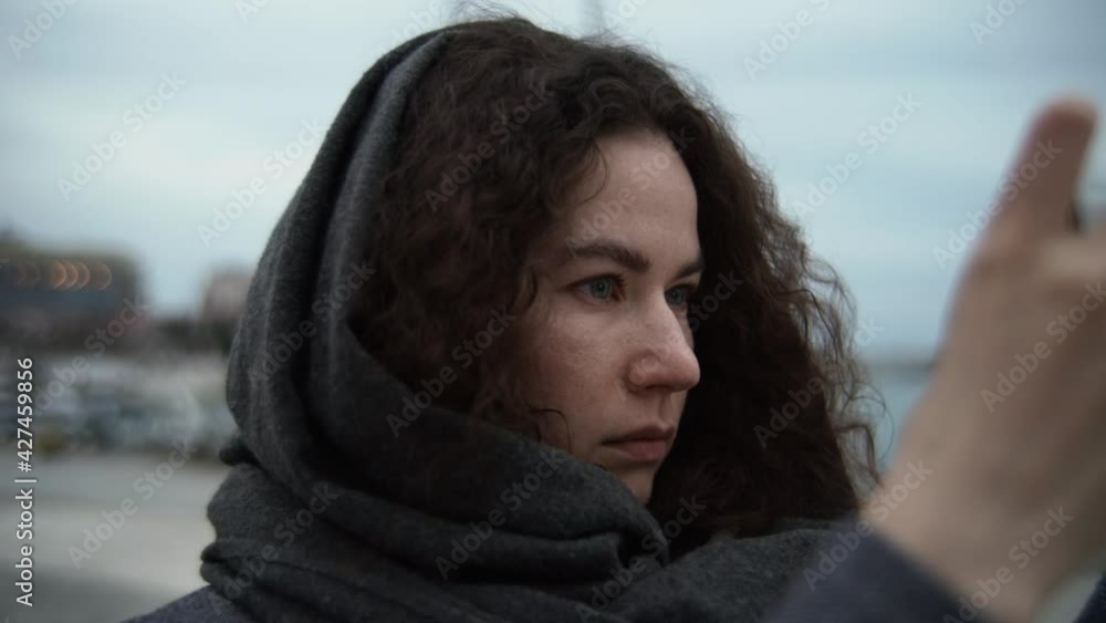 Young woman in a gray scarf with curly hair on a city street in the evening. holds a device in her hands and takes videos or photographs. The light from the screen illuminates the face. Young woman in a gray scarf with curly hair on a city street in the evening. holds a device in her hands and takes videos or photographs. The light from the screen illuminates the face.