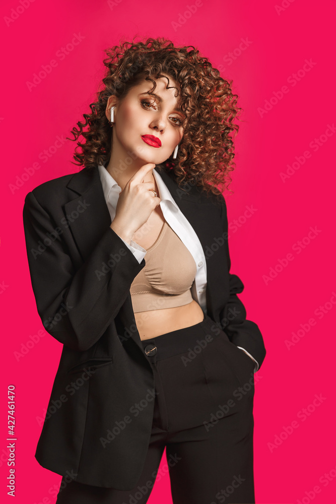 beautiful curly girl in an office black and white outfit listens to music on headphones. looks into the camera. isolated on bright pink background beautiful curly girl in an office black and white outfit listens to music on headphones. looks into the camera. isolated on bright pink background