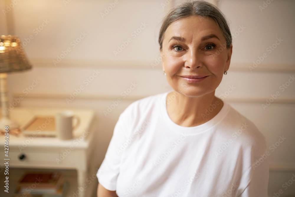 Mature people, aging, health and skin care concept. Portrait of beautiful positive middle aged female with wrinkles and gray hair posing indoors wearing white t-shirt, looking at camera with smile Mature people, aging, health and skin care concept. Portrait of beautiful positive middle aged female with wrinkles and gray hair posing indoors wearing white t-shirt, looking at camera with smile