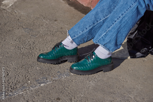 Women's feet in fashionable green shoes stand on the asphalt on a sunny day.