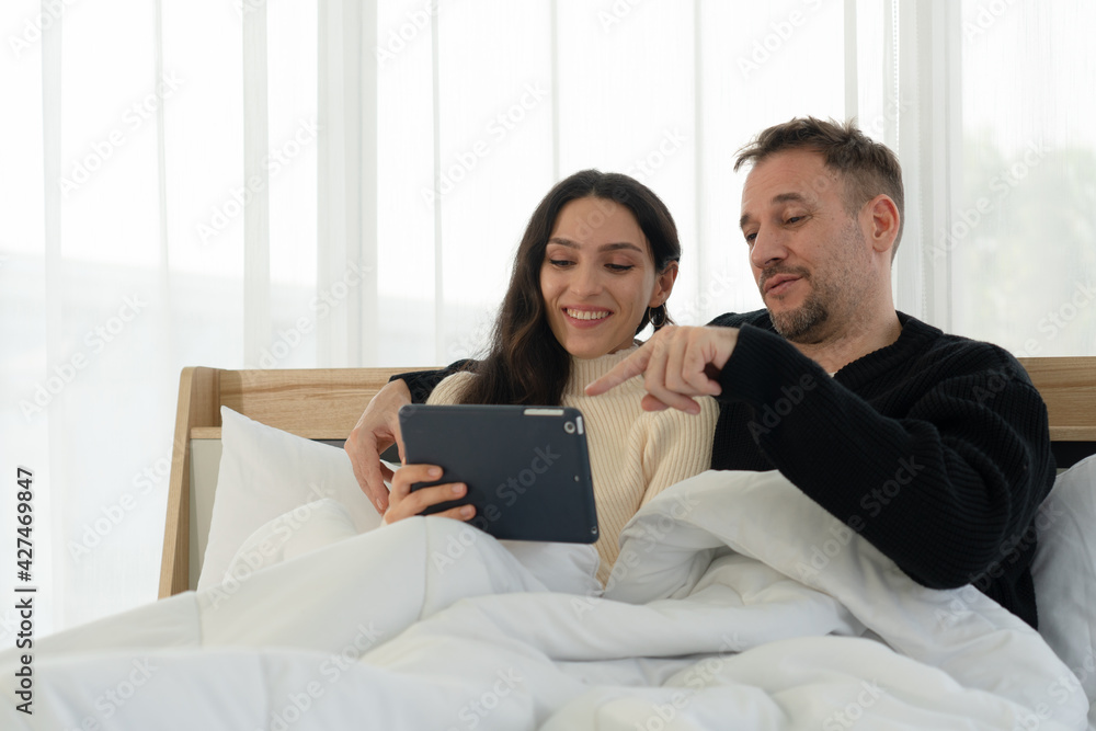 caucasian couple sitting in bed and watching movies from tablet in bedroom at home. togetherness concept