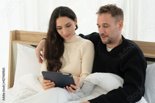 caucasian couple sitting in bed and watching movies from tablet in bedroom at home. togetherness concept © plo
