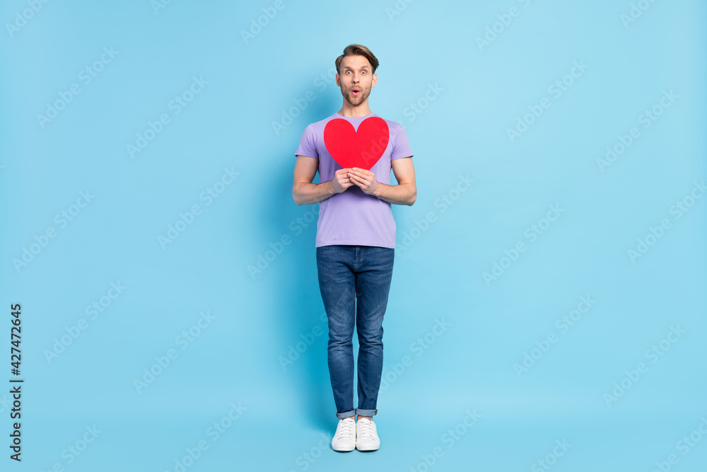 Full body photo of impressed positive young man hands hold heart symbol open mouth isolated on blue color background Full body photo of impressed positive young man hands hold heart symbol open mouth isolated on blue color background
