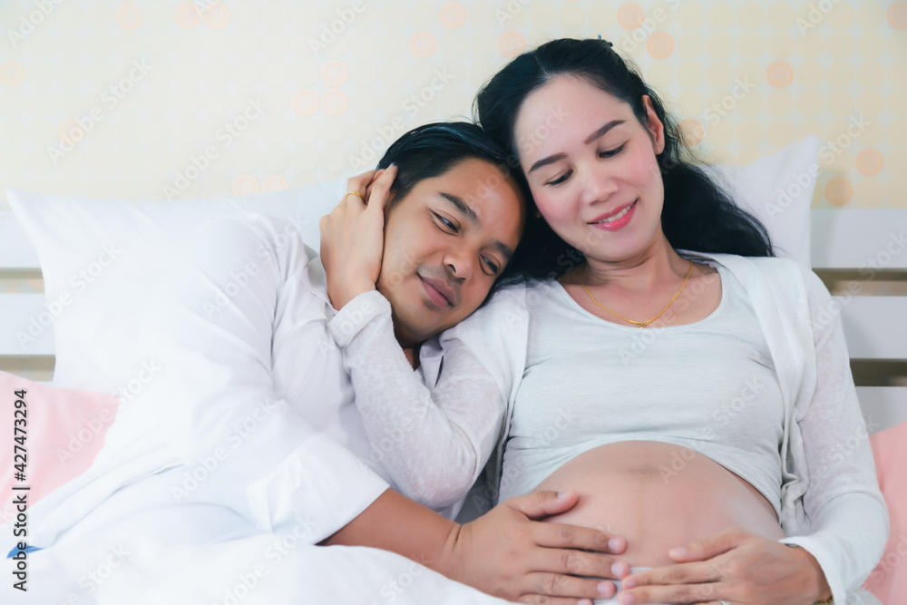 Asian Couple with Pregnant mother relaxing on bed together with love Loving man hug pregnant tummy wife at home Young couple expecting baby sit on bed together at bedroom Happy family Pregnancy mother