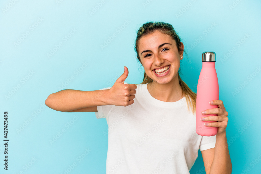 Young blonde caucasian woman holding a thermo isolated on blue background smiling and raising thumb up Young blonde caucasian woman holding a thermo isolated on blue background smiling and raising thumb up
