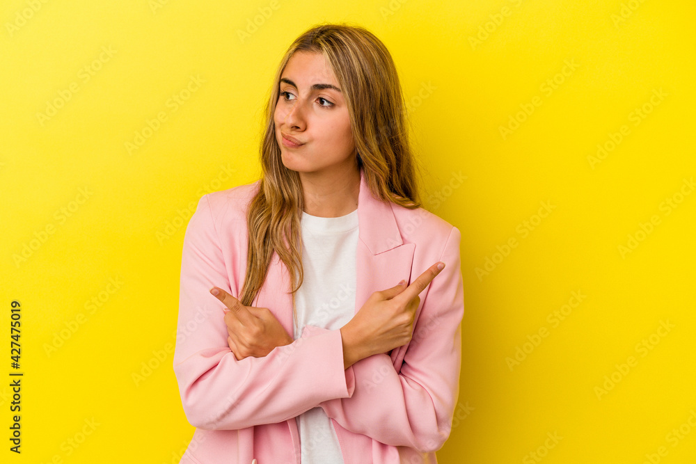 Young blonde caucasian woman isolated on yellow background points sideways, is trying to choose between two options. Young blonde caucasian woman isolated on yellow background points sideways, is trying to choose between two options.