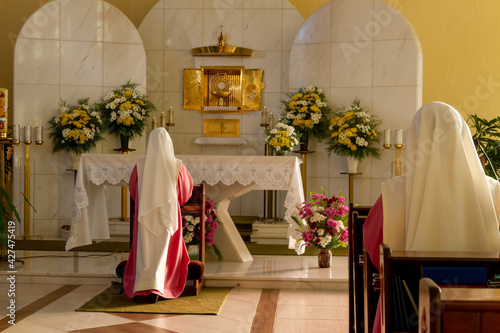 Photography Two nuns dressed in pink and white robes kneeling praying and worshiping the Blessed Sacrament of the Eucharist in a modern chapel with a flowered altar